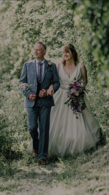 Bride and Groom walk towards the camera. She has a cascading bouquet of purple flowers.