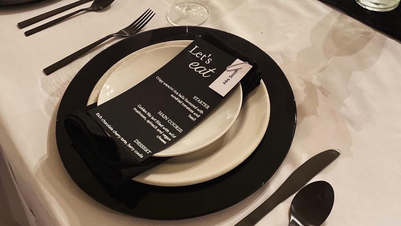 event styling image. Black charger plate with a white main plate and side plate on top. Black menu and cutlery on there.