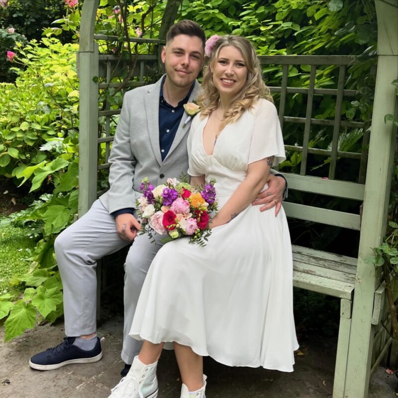 Beth and Alex (Bride and Groom) sit in the garden of their registry office. She holds a colourful bouquet and he has a white buttonhole.