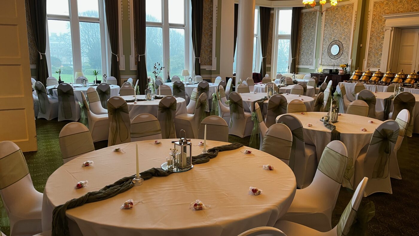 An event set up in a venue. Round tables with white table clothes, green sashes on the chairs and tables.