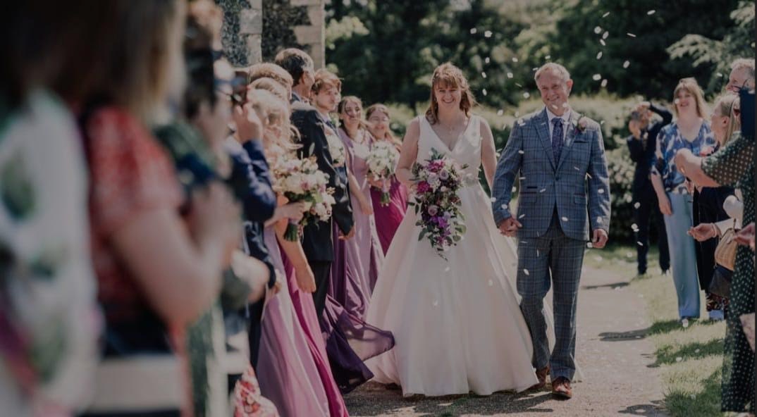 A bride and groom walk down their confetti line, she is holding a cascade bouquet and he has a buttonhole in the traditional style. Their colour palette contains lilac's and purples